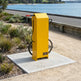 A bright yellow portable hydrogen fuel station mounted on a square concrete pad beside a sandy path and waterfront, with a coiled black hose and a small visible Paccase label on the front.