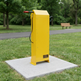 A bright yellow outdoor fitness machine mounted on a square concrete pad in a grassy park, with trees and a bench blurred in the background.