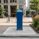 A blue portable electric vehicle charging station with a black cable,  placed on a concrete pad in a city plaza with a historic building in the background.