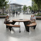 A modern outdoor round picnic-style table with integrated curved wooden benches and a seating area, shown in a public plaza. Two people are seated at the table, with others walking in the background near trees, benches, and a glass-front building.