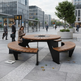A modern outdoor round fire pit table with curved wooden benches in a city plaza. A woman sits on the left bench, looking at her phone, while people walk in the background between tall glass office buildings.