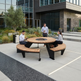 A modern round outdoor patio table with curved bench seating and a central fire pit, shown in a real-world outdoor patio setting with three people gathered around it.