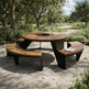 Outdoor round wooden fire pit table with curved wooden benches on a stone patio in a landscaped garden setting.