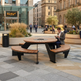 A modern round outdoor patio dining table with integrated curved wooden benches, shown in a real urban plaza with people sitting and walking nearby. The table has a dark metal frame and a circular fire pit opening in the center, set on a paved plaza with buildings and planters in the background.