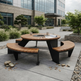 Outdoor round patio dining table with curved wooden benches and a black metal frame, set on a concrete plaza in front of a modern glass building.