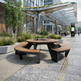 A modern outdoor dining set featuring a round wooden-top table with a dark metal frame and two curved bench seating pieces, placed in a landscaped urban walkway with plants and a glass-roofed structure in the background.