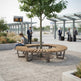 A curved wooden park bench arranged in a circle around a young tree at an outdoor transit station, with a woman sitting on the left side and several people standing nearby.