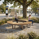 A curved wooden park bench with integrated seating is shown in a real outdoor plaza under a large tree, with two people sitting on it. The scene includes a bus stop, paved walkway, landscaping, and other park elements, so this is a Lifestyle image rather than a clean Product/Packaging shot.