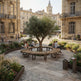 A circular wooden bench arrangement in a historic stone plaza, centered around a green tree, with people sitting and relaxing in an outdoor urban setting.