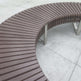 Curved outdoor bench with dark brown composite slats and stainless steel support posts on a light gray concrete surface.
