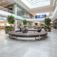 A large circular wooden bench with metal supports sits in the center of a modern shopping mall atrium with tiled floors, glass railings, potted trees, and several people sitting and walking nearby.