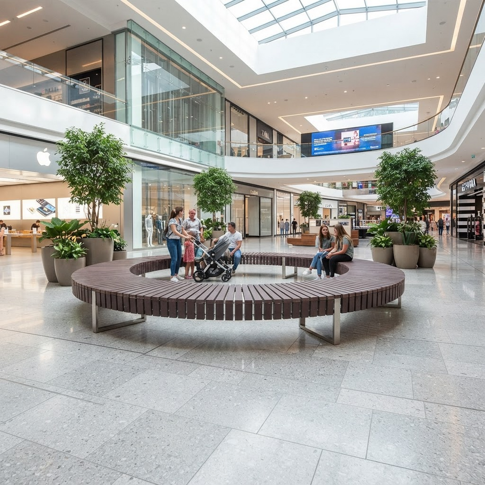 A large circular wooden bench with metal supports sits in the center of a modern shopping mall atrium with tiled floors, glass railings, potted trees, and several people sitting and walking nearby.