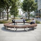 A large curved wooden bench with metal legs in an urban plaza, with several people sitting and reading or talking. Trees, buildings, and a city square are visible in the background.