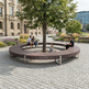 A large circular outdoor bench made of dark brown wood slats with a silver metal frame, arranged in a U-shape around a tree in a paved public square. Several people are sitting on the bench, including a child, in front of a grand classical building.