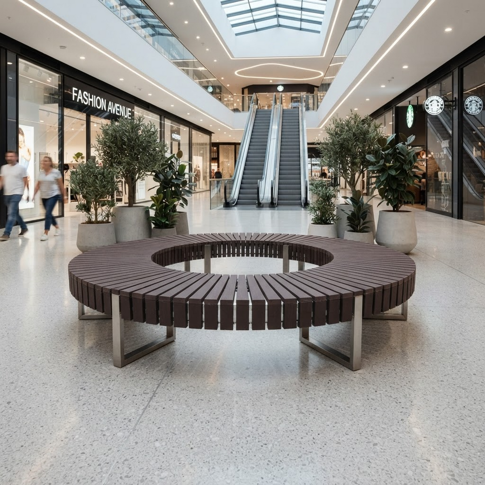 A round outdoor-style circular bench with a central opening, made of dark brown slats on a metal frame, situated in a modern shopping mall atrium with escalators and potted plants in the background.