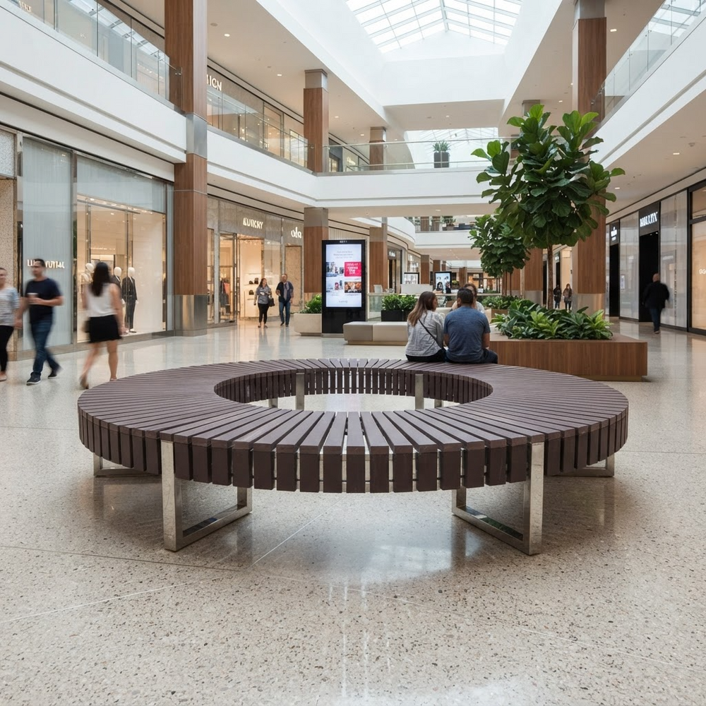 A large circular outdoor-style bench with dark brown slatted seating and silver metal supports, placed in a spacious modern shopping mall atrium. Several people are seated on the bench, and other shoppers walk nearby.