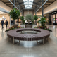 A large circular outdoor-style bench with dark brown slatted seating and silver metal supports, placed in a spacious indoor shopping mall corridor with potted plants, flowers, and visible shoppers in the background.
