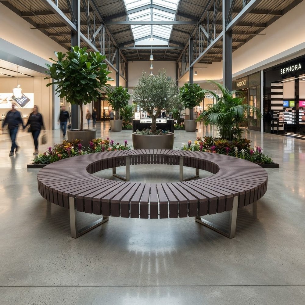 A large circular outdoor-style bench with dark brown slatted seating and silver metal supports, placed in a spacious indoor shopping mall corridor with potted plants, flowers, and visible shoppers in the background.