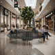 A modern curved black bench with a central planter holding a large green tree, situated in a bright, multi-level shopping mall atrium. Several people are sitting on the bench, and surrounding storefronts like LUNA and COSMOS are visible in the background.