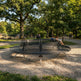 A large circular black metal park bench sits on a gravel-covered lawn in a sunny park, with people sitting on it and trees in the background.