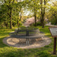 A modern circular metal fire pit bench with a slatted design sits on a gravel base in a grassy park area surrounded by green trees and a walking path in the background.