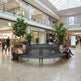 A large circular black slatted bench in a modern shopping mall atrium, with several people sitting on it. Visible storefronts include Zara, Starbucks Coffee, and Apple.