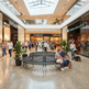 A wide view of a modern shopping mall atrium with multiple levels, a large curved black bench in the foreground, several people sitting and walking, and storefronts including Starbucks Coffee, a bookstore, and fashion retailers.