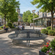 A modern circular metal park bench in a paved town square, surrounded by trees, flowers, and people sitting and walking in the background.