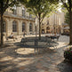 A modern circular bench made of dark metal slats sits in the center of a sunlit urban plaza with stone paving, trees, and people sitting on nearby benches. Historic beige buildings fill the background.