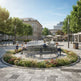 A modern outdoor plaza with a large circular bench surrounded by landscaped flowers and grass. Several people are seated on the bench, and the plaza is framed by trees and historic-style buildings under a clear blue sky.