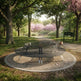 A modern circular metal park bench sits on a round concrete pad in a tree-lined park path with pink blossoms in the background.