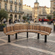 A curved wooden outdoor bench with black metal legs situated in a European-style town square with a fountain, historic buildings, and a red tram in the background.