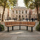 A curved wooden park bench with black metal legs situated in a paved public square, with historic buildings, trees, and small groups of people walking in the background.