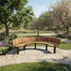 A curved wooden park bench with dark metal legs situated on a stone pathway in a lush, green park with blooming trees and a distant person sitting on a bench.
