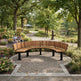 A curved wooden park bench with black metal legs sits on a paved circular walkway in a lush garden. In the background, a couple sits on a bench near flowers, with trees, a path, and a pond visible.