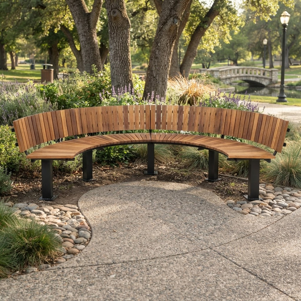 A curved wooden park bench with black metal legs, situated outdoors on a concrete path in a landscaped park with trees, shrubs, and a stone bridge in the background.