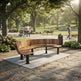 A curved wooden park bench with black metal legs sits on a concrete pad in a sunlit public park. Trees, walking paths, benches, people, and a trash can are visible in the background.