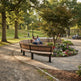 A curved wooden park bench with black metal supports is situated in a landscaped garden bed with flowers, surrounded by a paved path and trees in a park setting.
