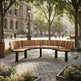A curved wooden outdoor bench with black metal legs situated in a paved urban plaza surrounded by trees, street lamps, and distant pedestrians.