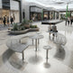 Modern outdoor plaza seating with curved metal benches, round metal tables, and matching stools in a landscaped shopping-center walkway. Several people are seated and walking nearby, with storefronts, plants, and a covered walkway visible in the background.