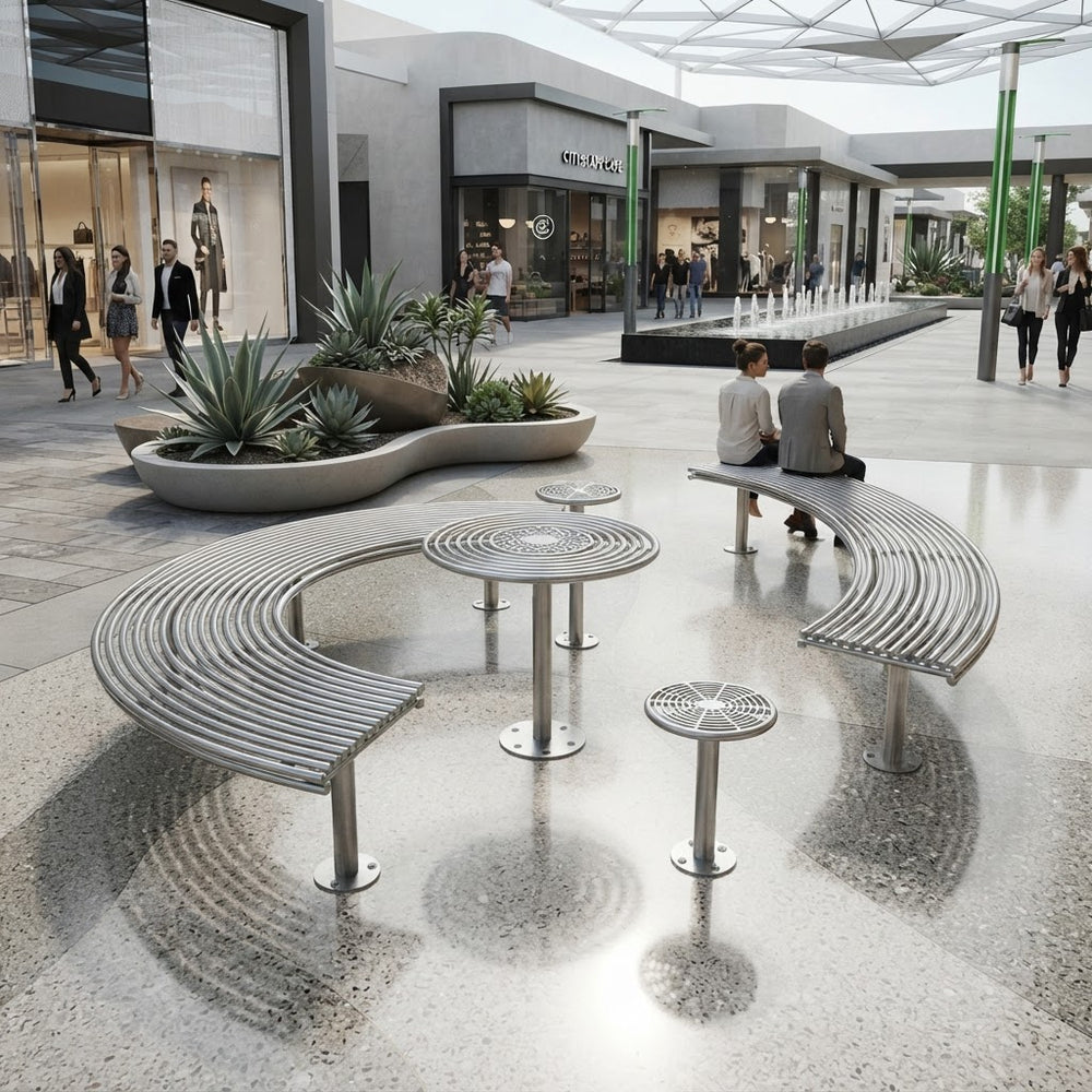 Modern outdoor plaza seating with curved metal benches, round metal tables, and matching stools in a landscaped shopping-center walkway. Several people are seated and walking nearby, with storefronts, plants, and a covered walkway visible in the background.