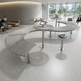 A modern curved metal lounge seating system with integrated round tables in a bright, contemporary office lobby. The furniture features a slatted silver design with circular floor mounts and central table bases, with a few people seated in the background near large windows.