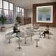 A modern arrivals lounge interior with curved metal tables and matching benches, several people seated and working or using a phone, large windows with a city skyline, and a wood-paneled wall with a illuminated sign reading 'RESERVATION SERVICES - ARRIVALS LOUNGE'.