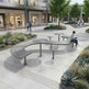 Curved modern metal outdoor seating with integrated tables in a landscaped plaza, with a woman sitting on one bench reading and shoppers walking in the background.