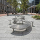 A modern curved stainless steel outdoor seating bench with matching small stools in a paved urban plaza surrounded by trees and buildings.