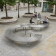 A modern curved stainless steel outdoor seating and dining system with integrated tables, shown in a public plaza with people sitting and walking nearby.