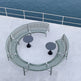 A curved outdoor seating set on a concrete deck overlooking the ocean, featuring a semi-circular metal bench with green slats, two round black tables, and a white railing above.