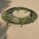 An olive green circular outdoor bench with a slatted metal design, shown from an elevated angle on a sandy ground.