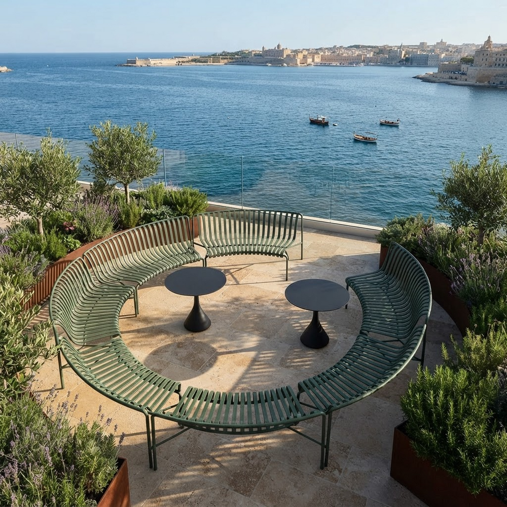 A curved green metal patio seating set with two small round black tables, placed on a stone terrace overlooking a blue sea and coastal city.