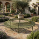 An olive green curved metal garden bench with a slatted seat and back, situated in a luxurious Mediterranean-style courtyard with stone paving, potted olive trees, and purple lavender flowers.